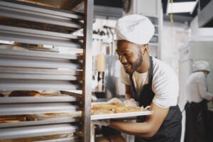 A cheerful baker in a professional kitchen holding a tray of freshly baked breads. - Photo by Gustavo Fring on Pexels