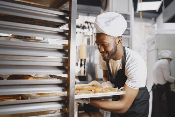 A cheerful baker in a professional kitchen holding a tray of freshly baked breads. - Photo by Gustavo Fring on Pexels