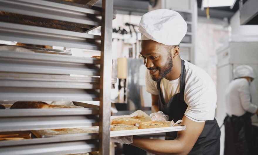 A cheerful baker in a professional kitchen holding a tray of freshly baked breads. - Photo by Gustavo Fring on Pexels